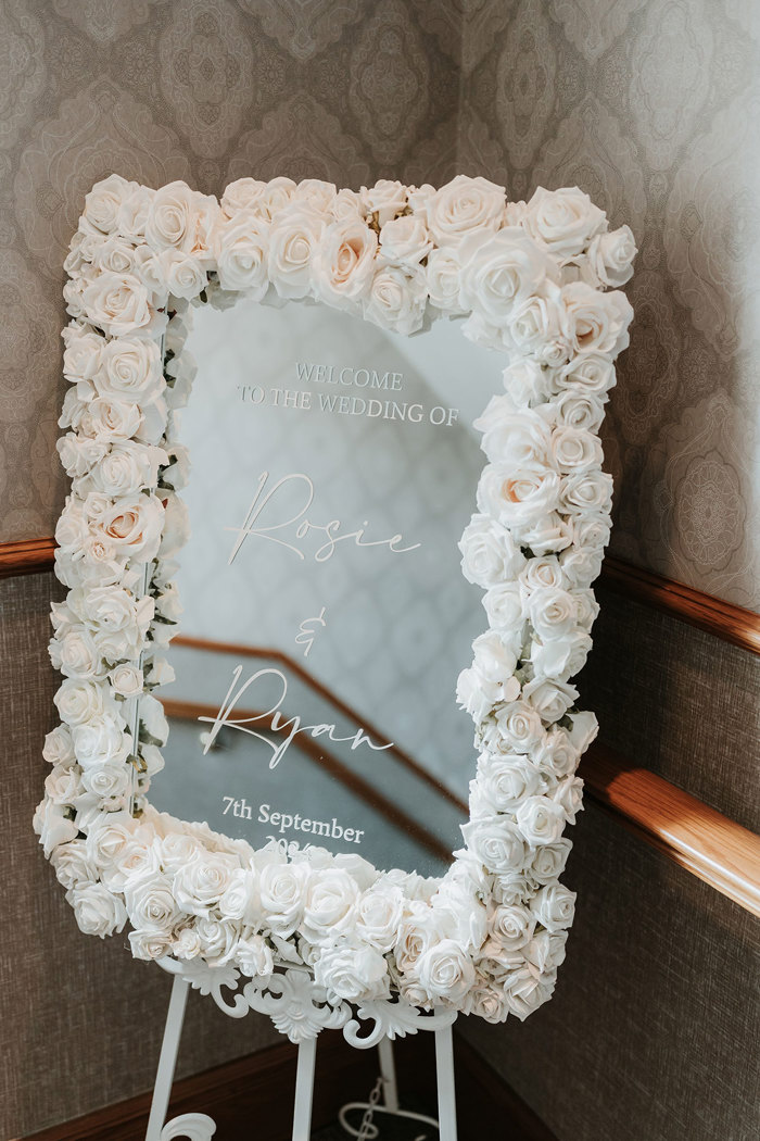 a mirrored wedding welcome sign framed by lots of white roses 