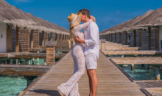 a woman in a white outfit and bride embroidered sun hat kisses man in white outfit on above-water villa boardwalk