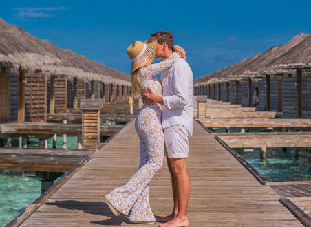 a woman in a white outfit and bride embroidered sun hat kisses man in white outfit on above-water villa boardwalk