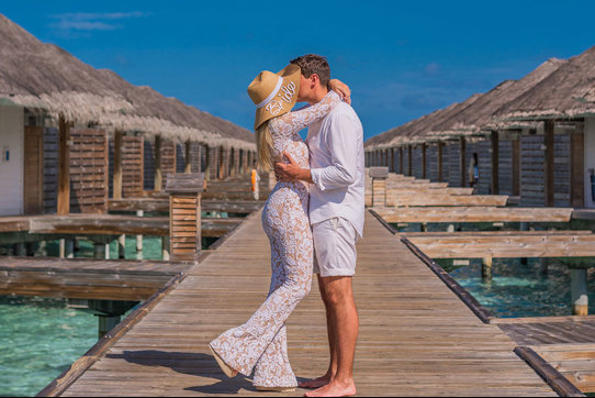 a woman in a white outfit and bride embroidered sun hat kisses man in white outfit on above-water villa boardwalk