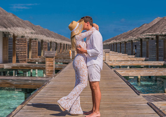 a woman in a white outfit and bride embroidered sun hat kisses man in white outfit on above-water villa boardwalk