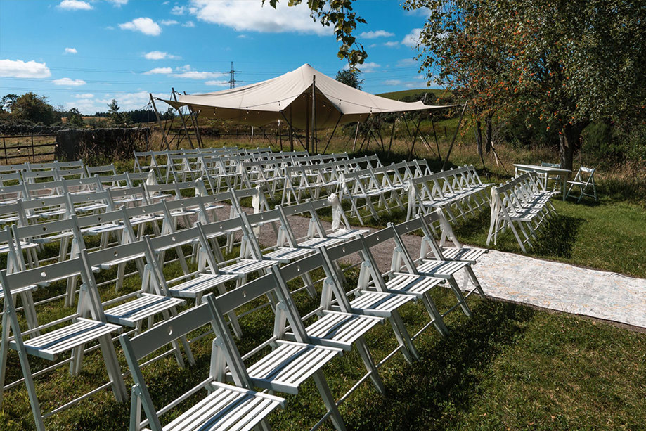 Rows of white wooden chairs set for a ceremony outside with a small stretch tend behind it