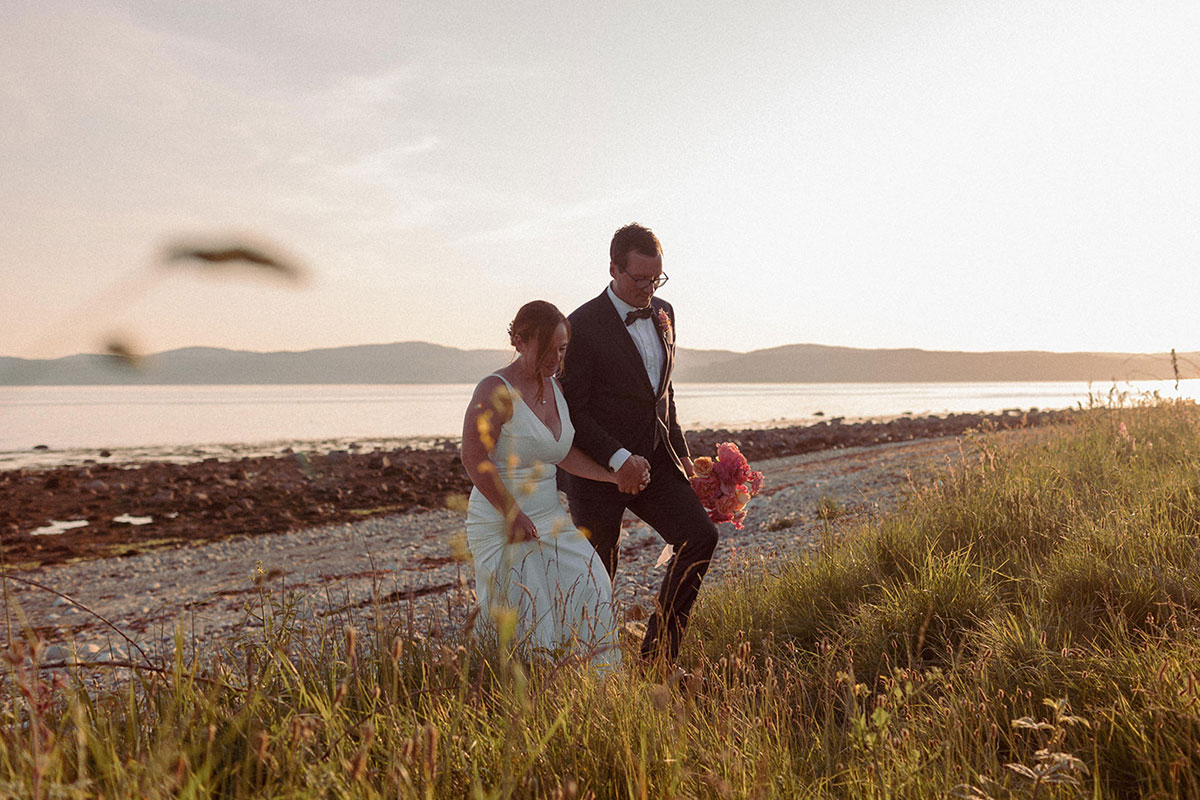 bride in strappy wedding dress and groom in black tuxedo hold hands as they walk away from sandy beach at sunset, starting to step through tall grass