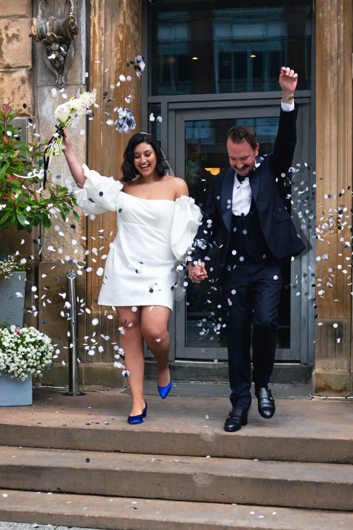 A bride in a short wedding dress and a groom in a navy tuxedo have white confetti thrown over them as they walk down stone stairs