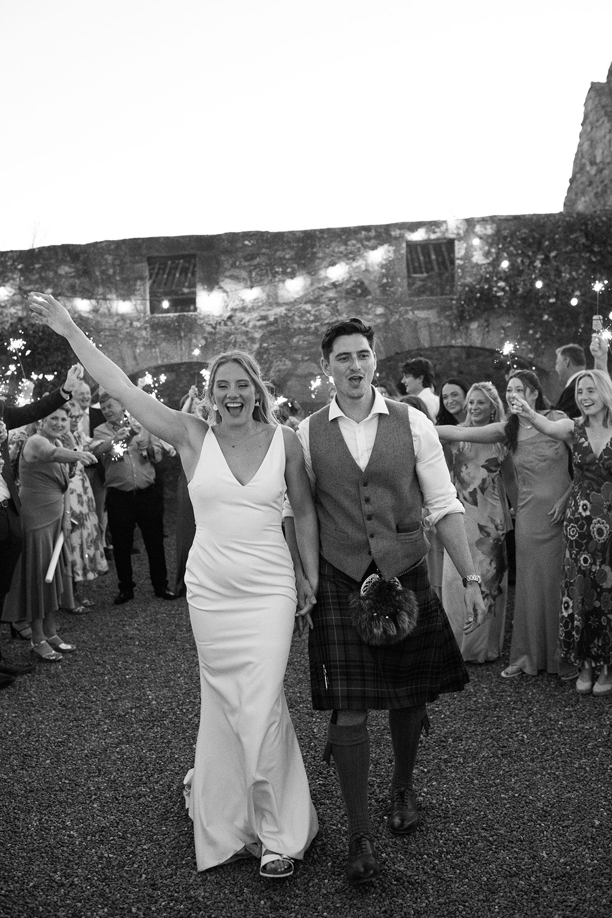 Bride and groom walk hand-in-hand during their sparkler exit, smiling as guests form a tunnel of lights behind them