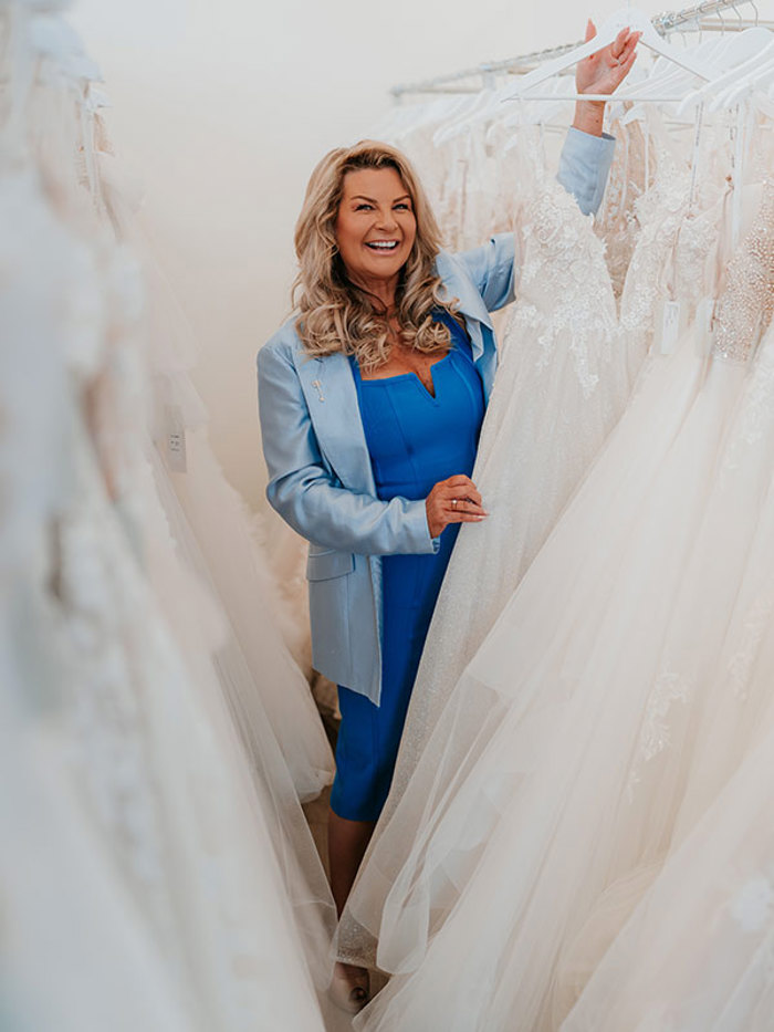 a blonde woman in a bright blue suit selecting a hanging wedding dress from full display rails