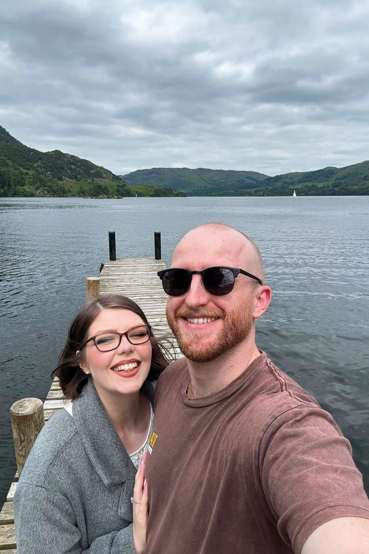 Couple taking a lakeside selfie on a wooden jetty overlooking a scenic Lake District landscape with rolling hills and calm water