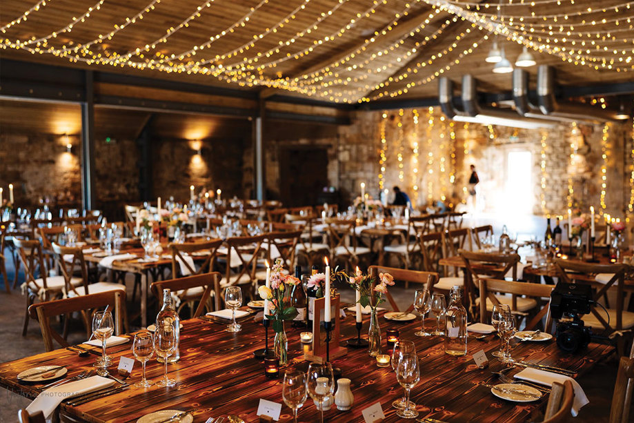 Inside a large barn with wooden tables, which have cutlery, candles and flowers on them, there is a canopy of fairylights on the ceiling