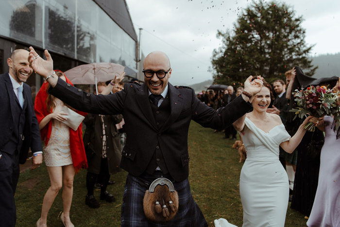 groom in blue, grey and white tartan kilt outfit walk with arms out with falling flower confetti around him and bride in off the shoulder white wedding dress holding a bouquet