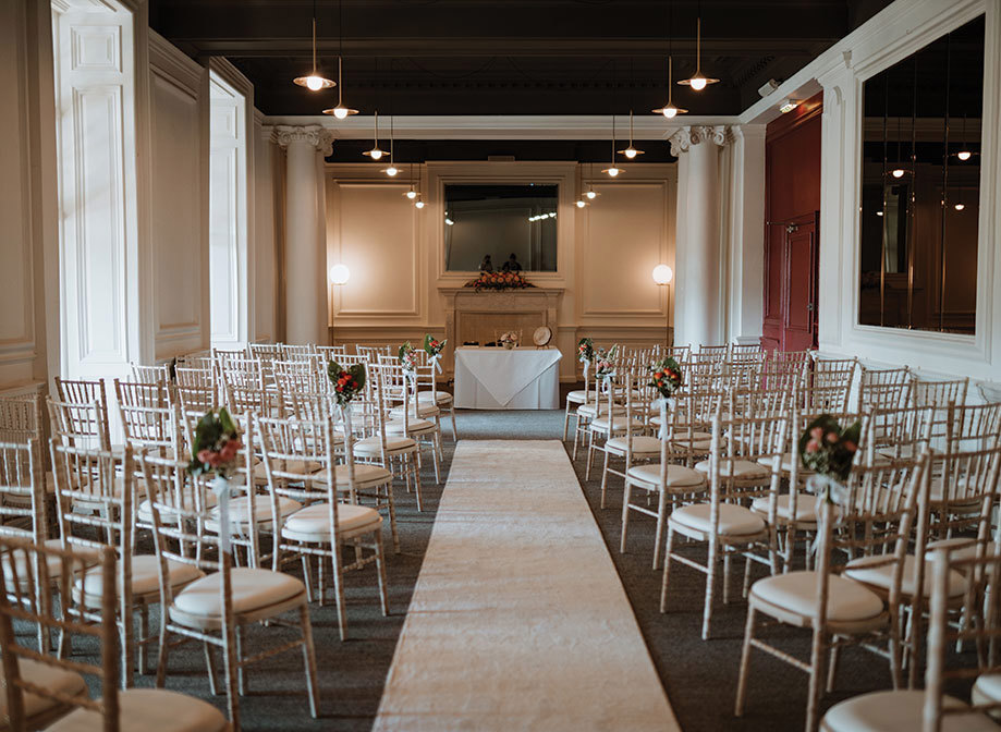 A long room with a white aisle runner in the centre, with white chairs set up on either side for a ceremony