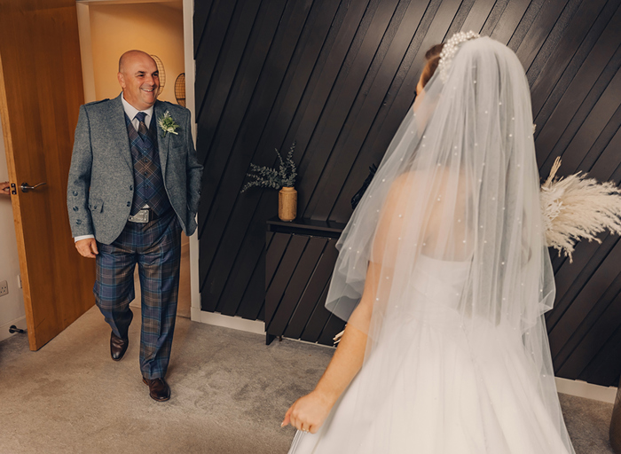 Man in highlandwear walking in and seeing bride and smiling