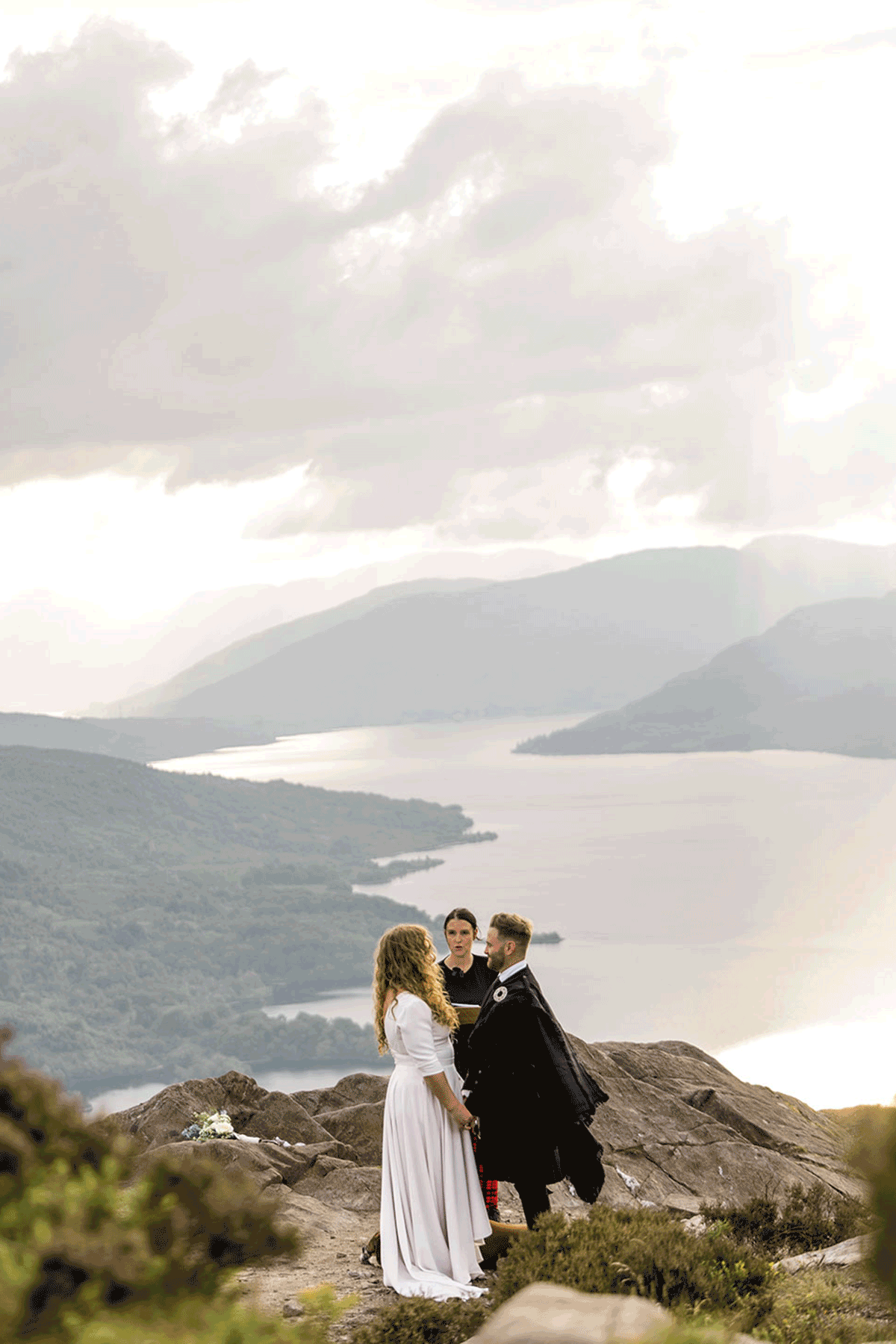 Couple exchanging vows during a Scottish mountain elopement overlooking Loch Lomond, with dramatic skies and panoramic Highland scenery behind them