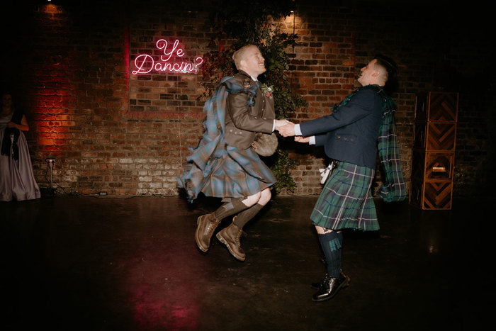 Two Men In Kilts Dancing With Ye Dancing Neon Sign And Red Brick Wall In Background