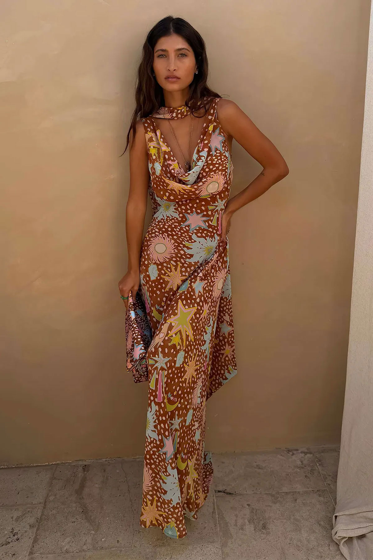 Brown printed maxi dress with star and abstract pattern, cowl neckline and matching scarf detail worn outdoors against a neutral wall