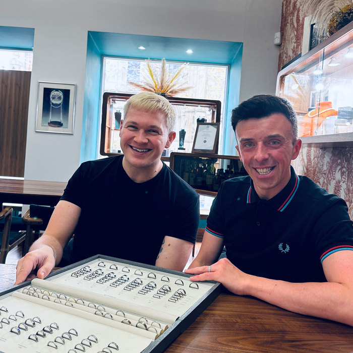 two men sitting behind a desk. There is a board of wedding rings in front of them on a wooden table.