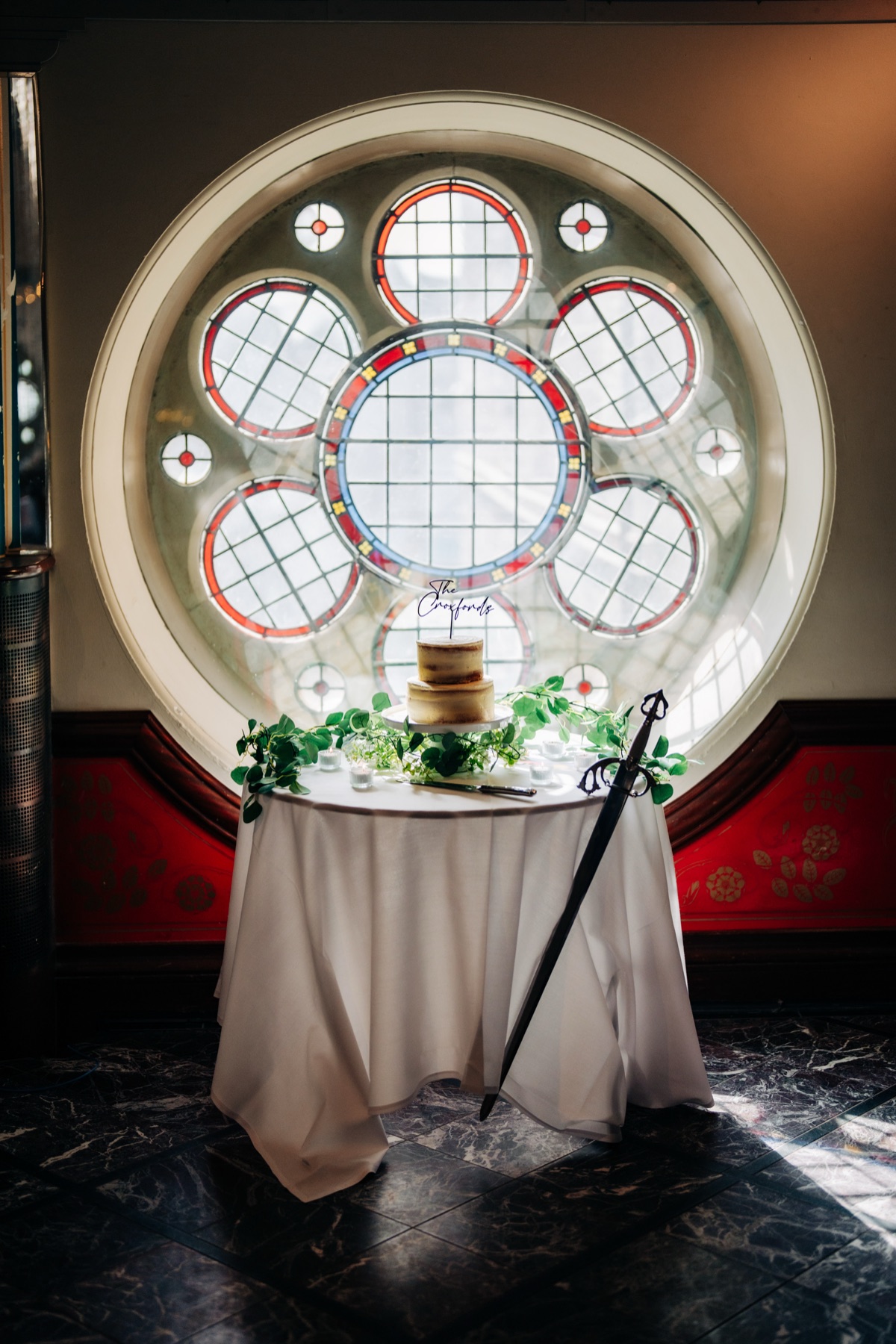 Two-tier wedding cake displayed in front of a circular stained-glass window at Òran Mór in Glasgow, surrounded by greenery and candles