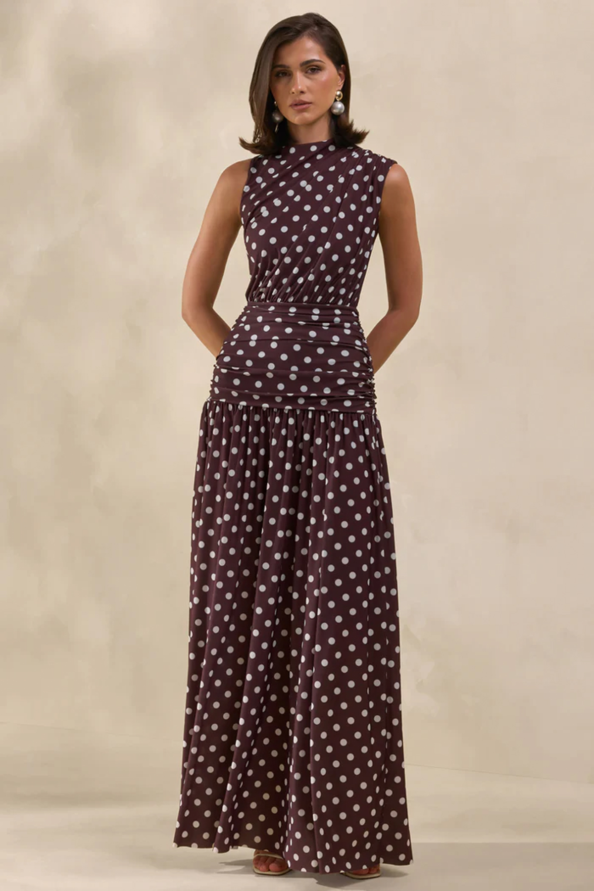 Woman wearing a brown and white polka-dot maxi dress with a high neckline, ruched waist and flowing skirt.