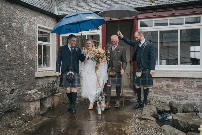 Guests cover the bride and her father with umbrellas as they leave their house