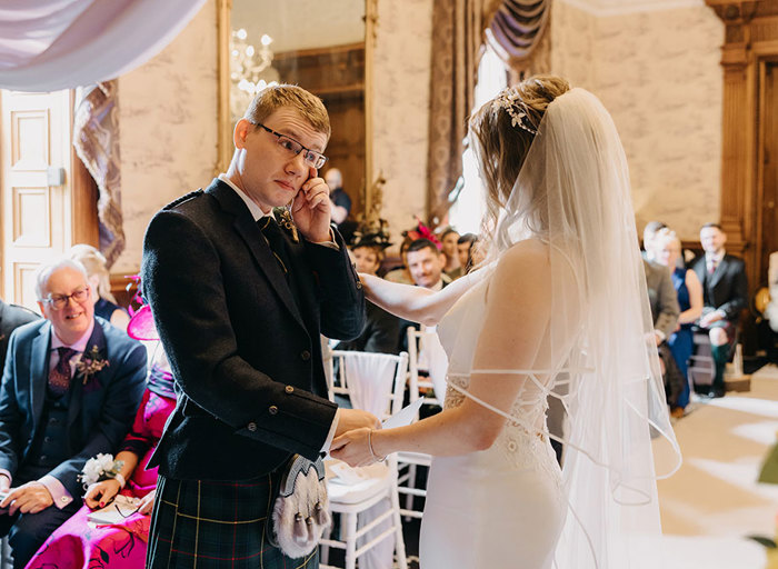 A groom wipes a tear from his eye during the wedding ceremony while holding hands with the bride