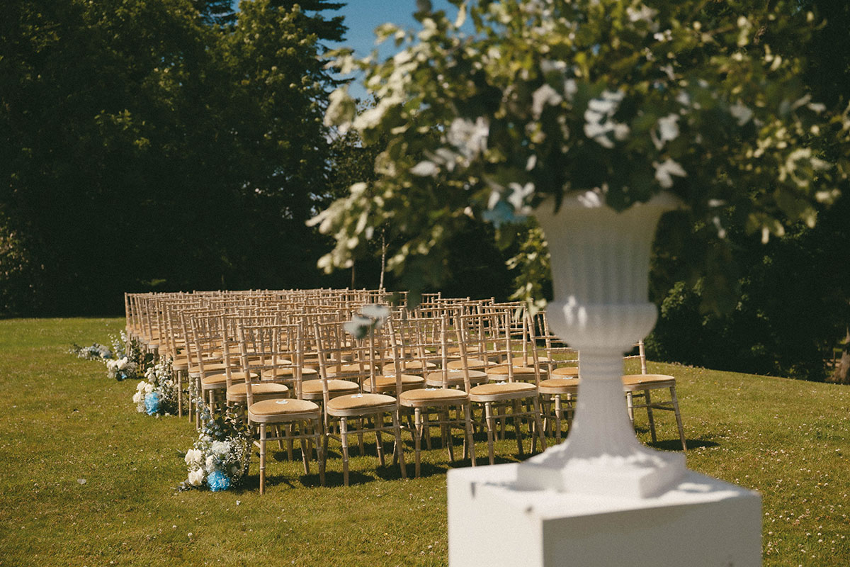 Outdoor wedding ceremony set up on the lawn at Aswanley, with rows of wooden chairs, blue and white aisle flowers and a white floral arrangement in the foreground
