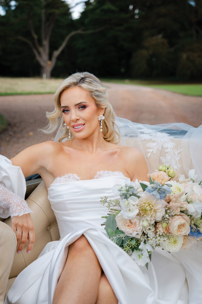 A bride in a sweetheart neckline dress with puffy sleeves and a slit in the skirt sits in a convertible car holding a bouquet of pastel flowers