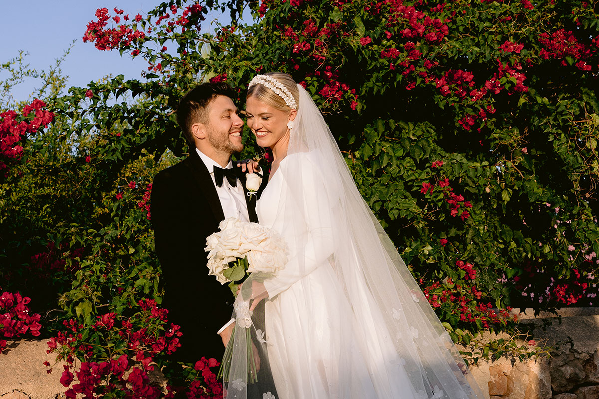 bride in long sleeve wedding dress with long floral applique veil and groom in tux lean into each other and smile in front of red flower tree
