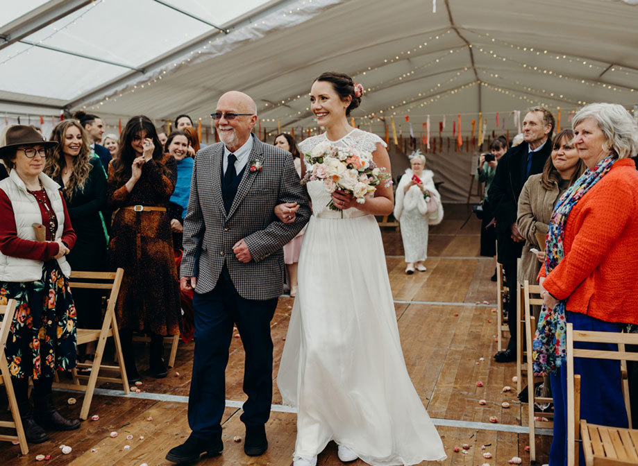 a bride walking down the aisle in a marquee on the arm of a man wearing a checked suit jacket. Guests are standing up from folding wooden chairs. The bride is wearing a lace top, flowing skirt and has colourful flowers in her hair