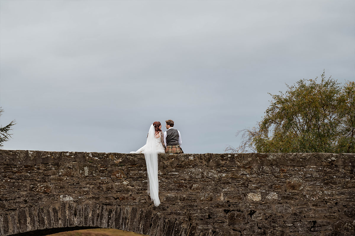 A bride and groom sit on the stone wall of a bridge with the bride's veil hanging over the edge