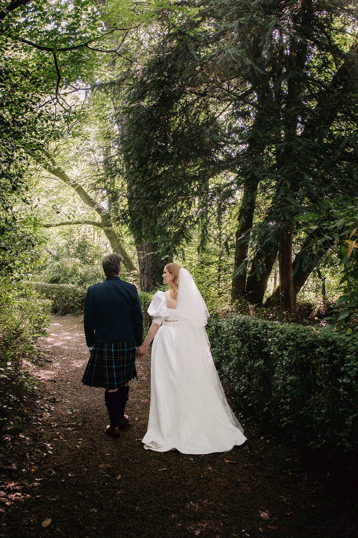 A bride and groom holding hands and walking along a tree-lined garden path at the Royal College of Physicians of Edinburgh, surrounded by greenery