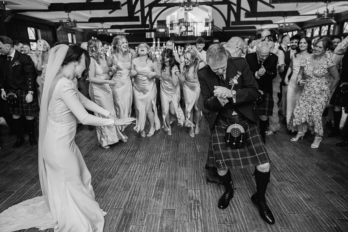 Bride dances with a kilted guest during a lively wedding reception, surrounded by cheering bridesmaids in satin dresses and guests clapping on the dance floor