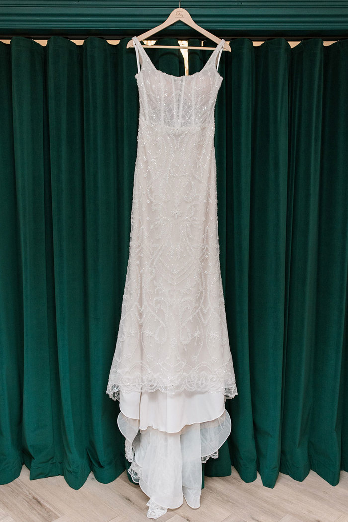 long beaded wedding dress is displayed on wooden hanger in front of dark green curtain