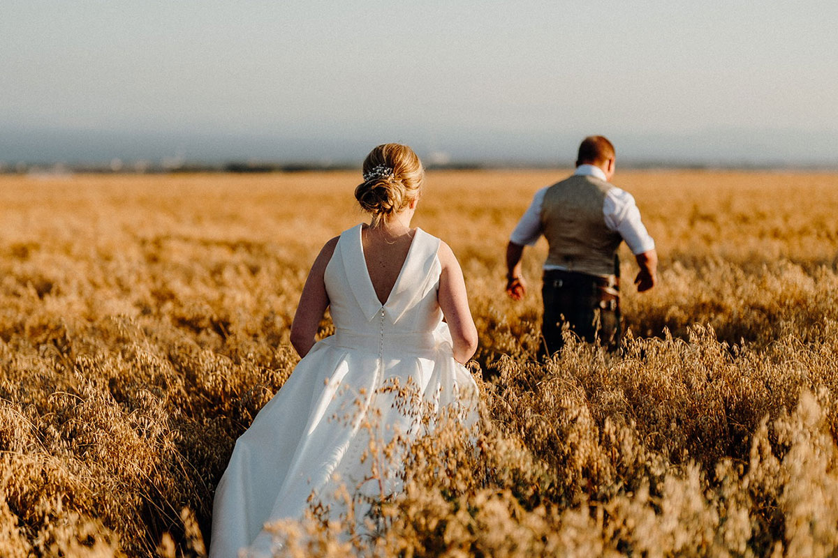 Bride and groom walking through a golden field at sunset during a Pratis Barns wedding in Fife, Scotland.