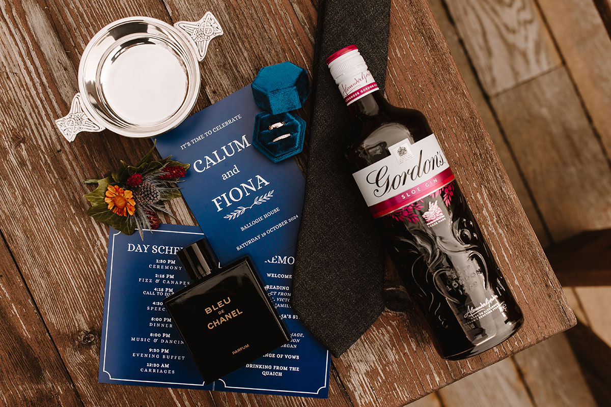 Wedding flatlay featuring invites, rings, quaich, Gordon’s sloe gin, Bleu de Chanel, tie, and buttonhole on wooden surface.