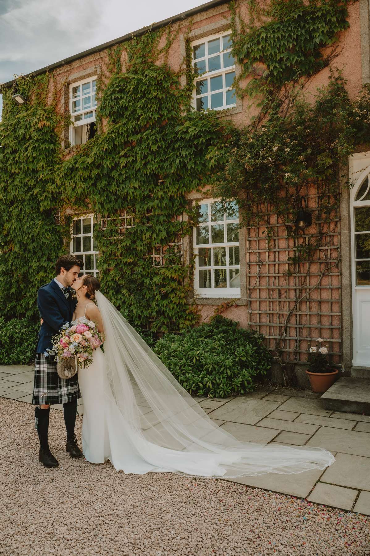 Bride and groom kiss beside the ivy-covered pink façade of Ballogie House
