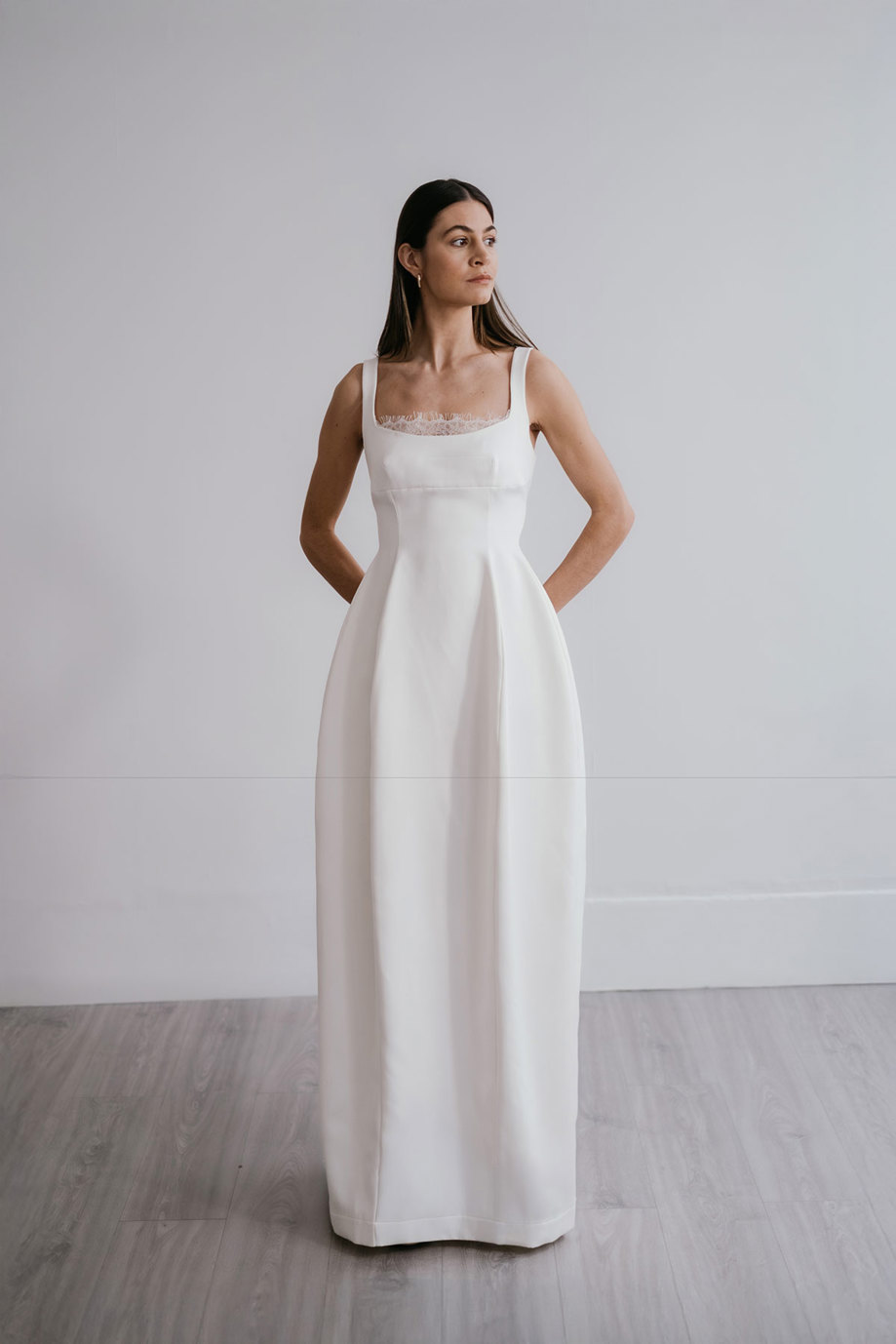 dark haired model stands in front of a plain white wall wearing a structured white wedding dress that has a border of lace peeking out of the bodice and an overall hourglass shape