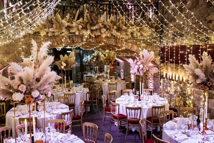 a bold and busy set up of round dinner tables with gold chars and golden stands of pampas grass florals centred at each table, strings of fairylights draping the entire room