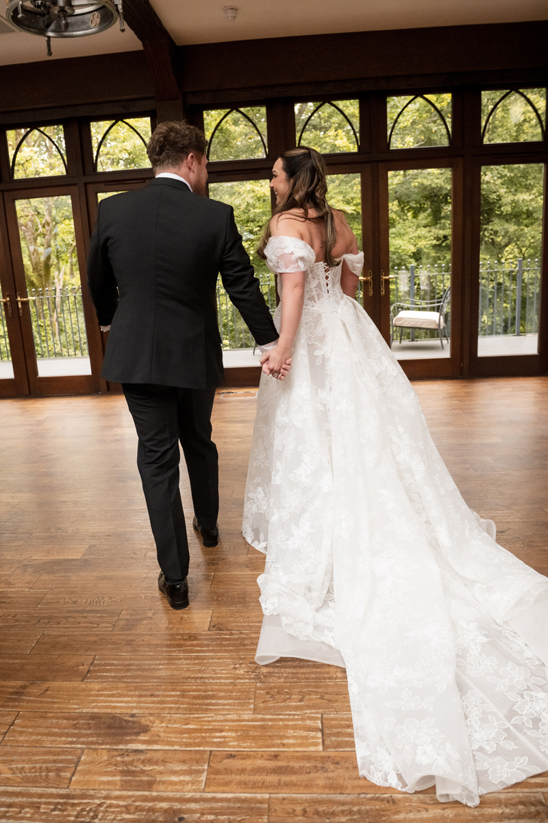 Bride and groom holding hands in the ballroom at Crossbasket Castle, showing the bride's floral lace wedding dress and train.