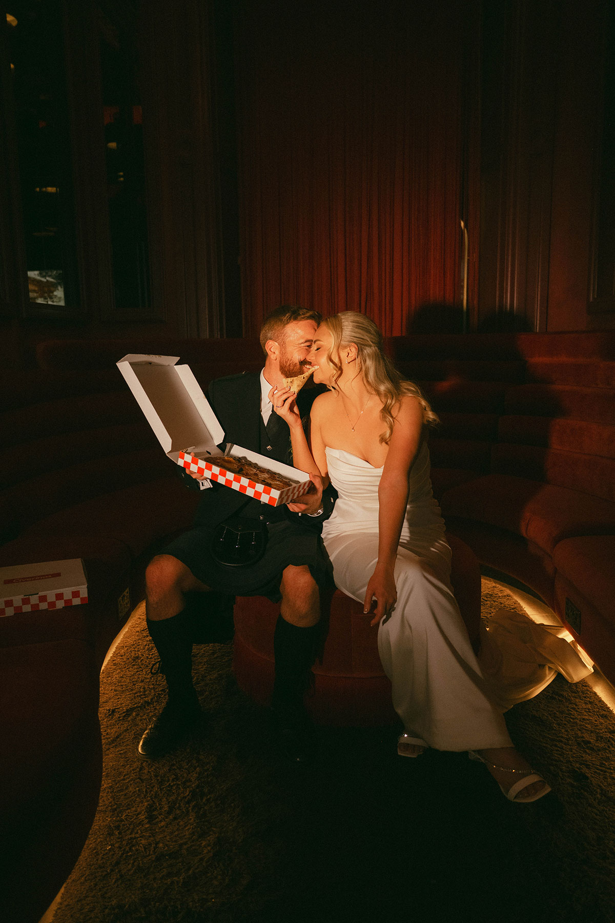 Image 2 bride and groom eating pizza on red velvet seating during relaxed wedding reception