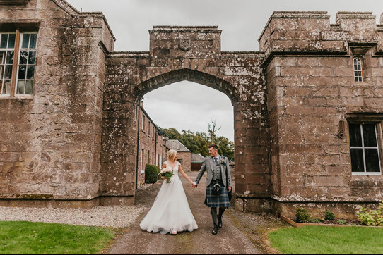 A bride in a long white dress and a groom in a blue and grey kilt hold hands and walk though a stone archway