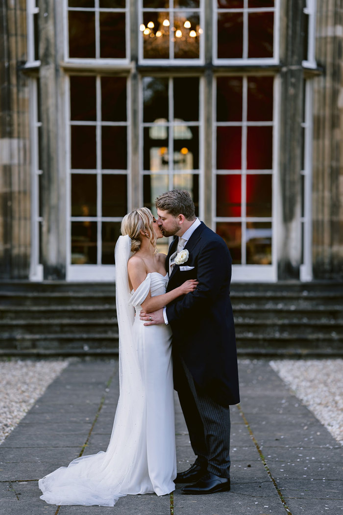 A bride and groom in wedding attire kissing in front of a large window and stairs at Mar Hall.