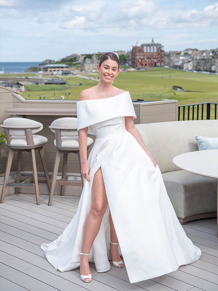 A bride standing in a rooftop bar with a golf course in the background wearing her hair in a bun and a white off-the-shoulder dress with a slit up on side and white strappy high heels