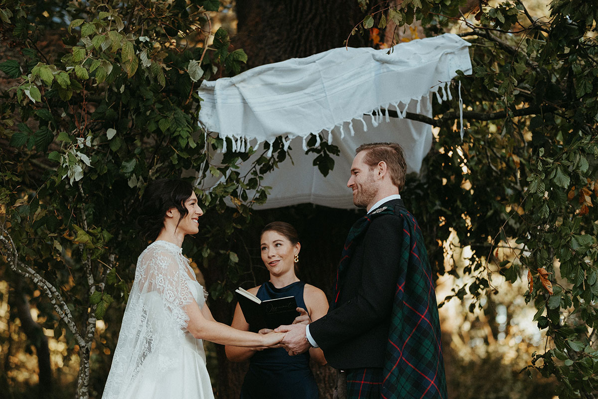 Outdoor wedding ceremony beneath a towering tree and chuppah at Cambo Country House, with guests gathered to witness the couple exchange vows.