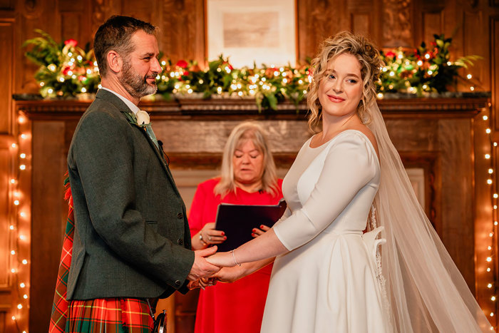 bride in white wedding dress and long veil and groom in red and green tartan kilt hold both hands out in front of them as a woman in front of them reads from a black folder