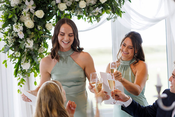 Two women in coordinating green dresses smiling and toasting with champagne at an Old Course Hotel wedding, under a floral arrangement