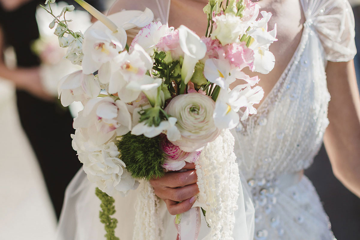 Bride in beaded gown holding a soft white and blush wedding bouquet featuring orchids, calla lilies and trailing amaranthus