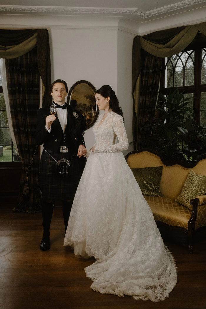 a bride wearing a lace dress and a groom wearing a black kilt outfit posing for a picture in a formal living room.