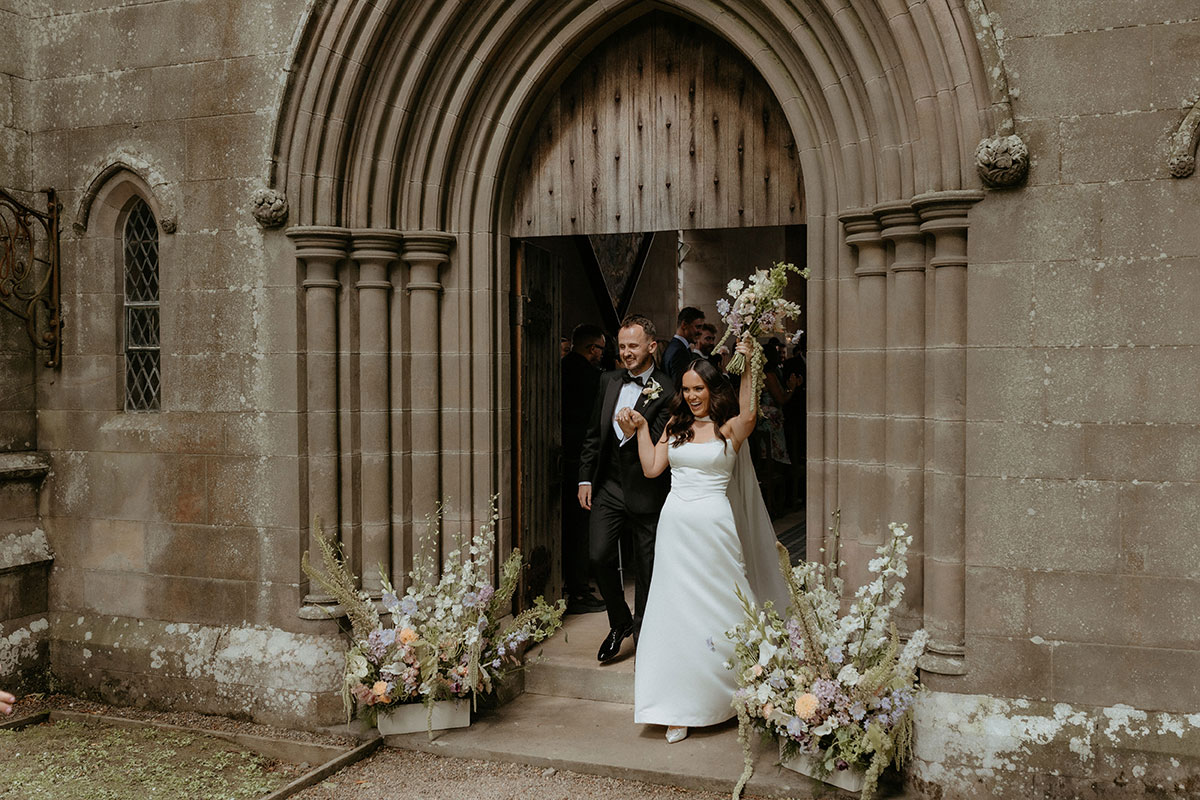 Bride and groom exiting Rosebery House chapel in Midlothian, celebrating with raised bouquet after ceremony.