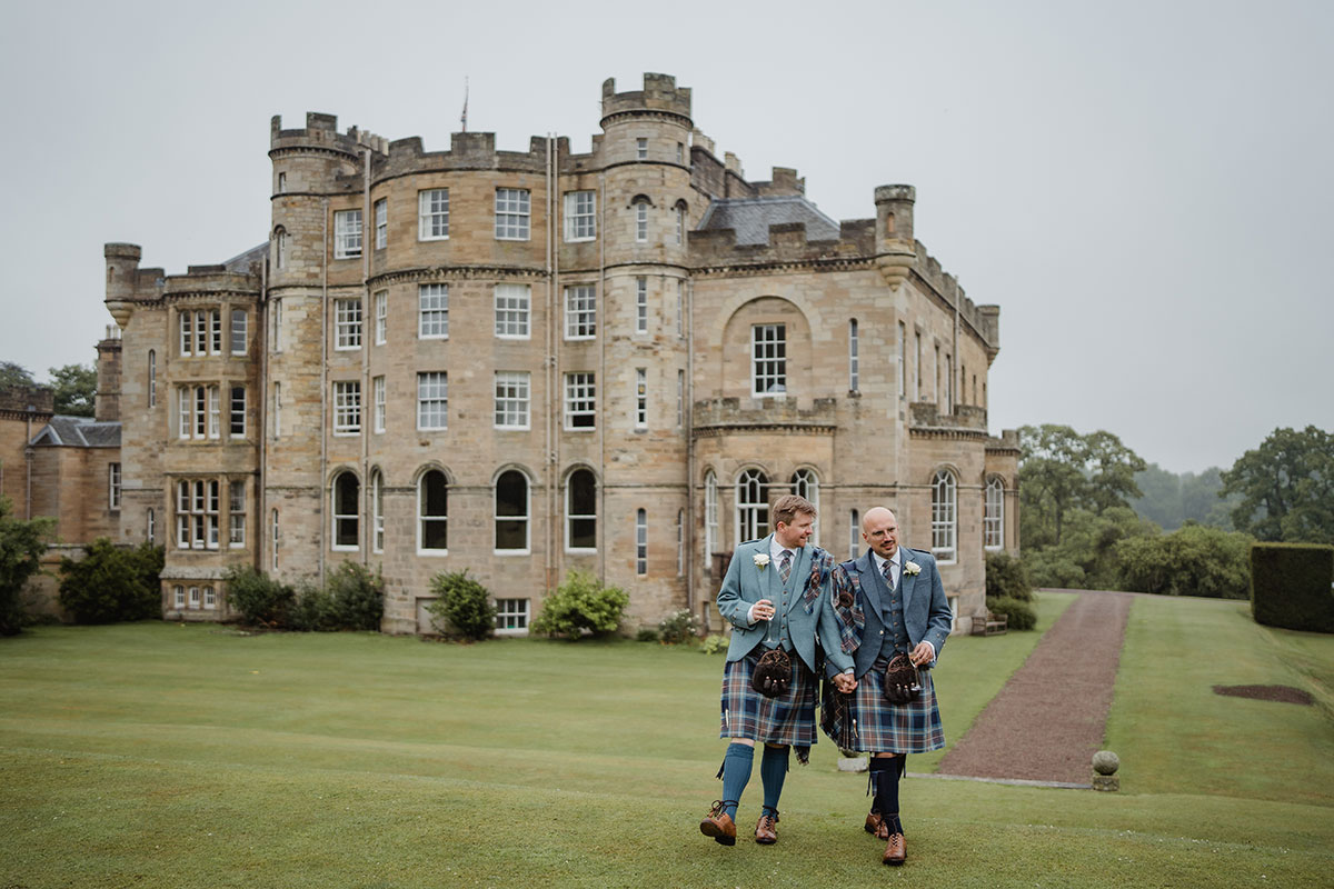 The newlyweds walk hand in hand across the lawn outside a historic stone castle, both wearing matching blue tartan kilt outfits