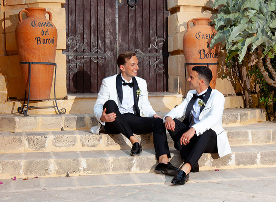 two grooms wearing white suit jackets and bow ties sitting on a set of stone steps in front of a large wooden door. There are two large terracotta urns reading 'Castello dei Baroni' either side