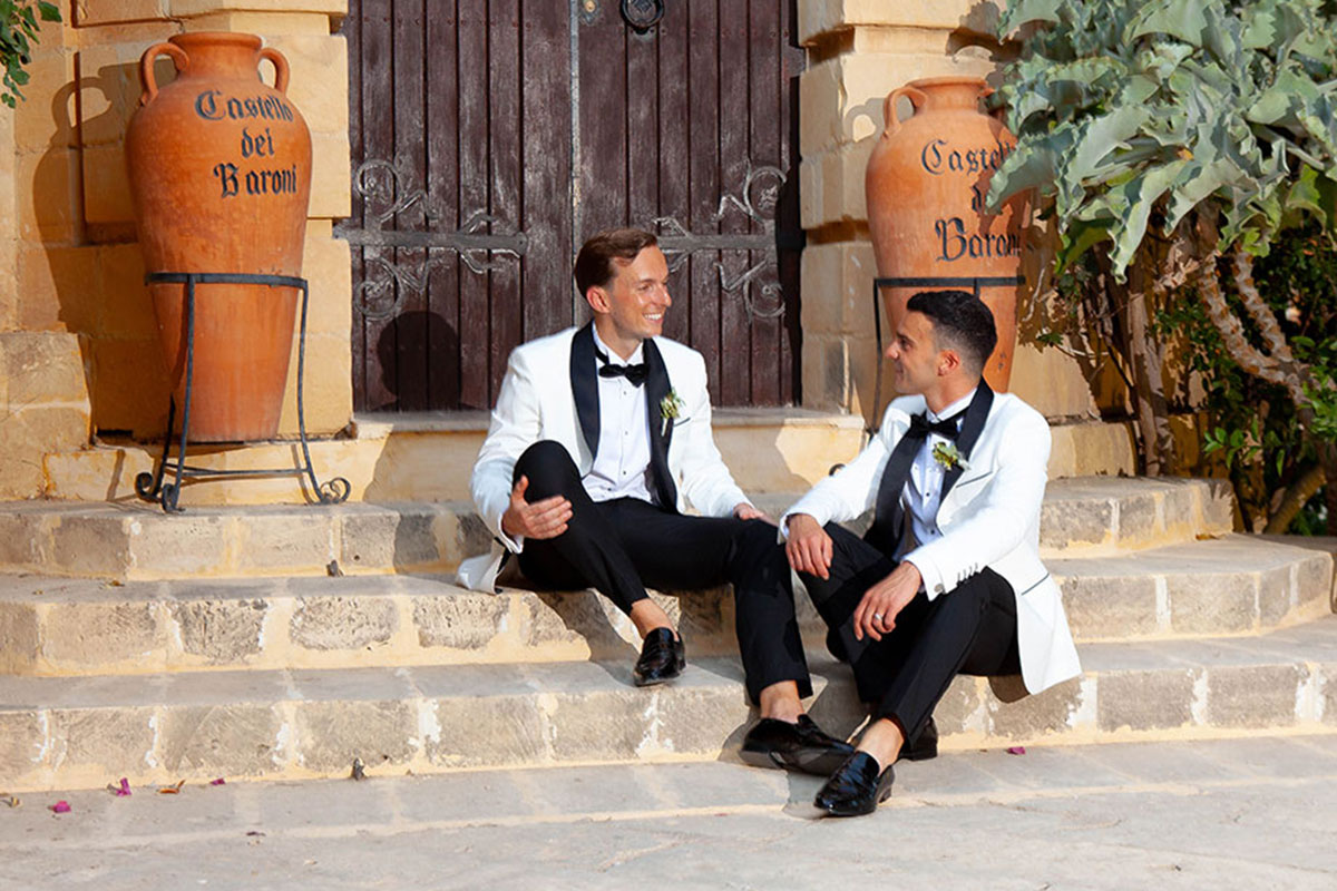 two grooms wearing white suit jackets and bow ties sitting on a set of stone steps in front of a large wooden door. There are two large terracotta urns reading 'Castello dei Baroni' either side